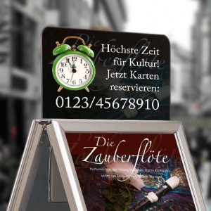 Aufmerksamkeitsstarke Top-Schilder stellen eine ideale Ergänzung zu Ihrem Kundenstopper dar! Im Bild ist vor einem verschwommenem Hintergrund einer Großstadtstraße ein vom Bildausschnitt angeschnittener Kundenstopper zu sehen, der mit einem Werbeplakat für "Die Zauberflöte" ausgestattet ist. Eine oben auf dem Kundenstopper angebrachtes Top-Schild verweist unter Zuhilfenahme eines Weckerbildes auf die Ticket-Hotline.