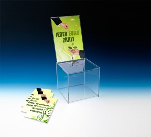 Spendenaktionen realisieren Sie professionell und unkompliziert mit abschließbaren Spendenboxen. Vor einem blauem Hintergrund ist eine Spendenbox mit integriertem Top-Schild platziert. In dieses ist ein Plakat eingeschoben, auf dem zwei Hände zu sehen sind. Beide Hände gehören offenbar zu einer Anzug tragenden Person, jedoch nicht zu der selben. Die eine Hand ist dabei, der anderen eine Münze zu geben. In der Hand des Empfängers ist bereits ein kleiner Stapel Münzen, aus welchem eine grüne Pflanze herauswächst. Beschriftet ist das Poster mit "Jeder Euro zählt". Neben der unbefüllten, transparenten Spendenbox liegen einige Flyer, die mit geicher Bildsprache arbeitet. Die Flyer sind mit folgendem Aufdruck versehen: "Wir setzen uns für blühende Aussichten weltweit ein".